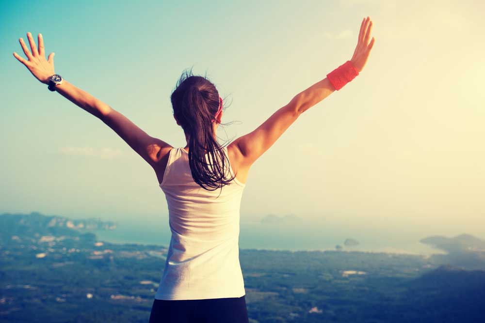 Woman raising both arms on a mountaintop overlooking the coast at sunrise